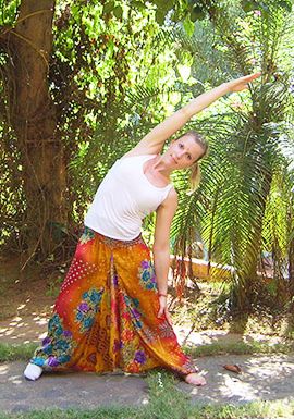 Dreieck Die Kursleiterin steht vor Palmen und macht eine stehende Yoga-Stellung, indem sie den rechten Arm nach schräg oben streckt und sich nach links beugt. Die Beine sind gespreizt und der linke Arm hängt nach unten. Diese Stellung nennt man Dreieck.